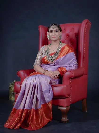 Seated gracefully, the model showcases the beautiful drape and color combination of the lavender and orange Kanchipuram style saree. This piece is a blend of tradition and modern color palettes.