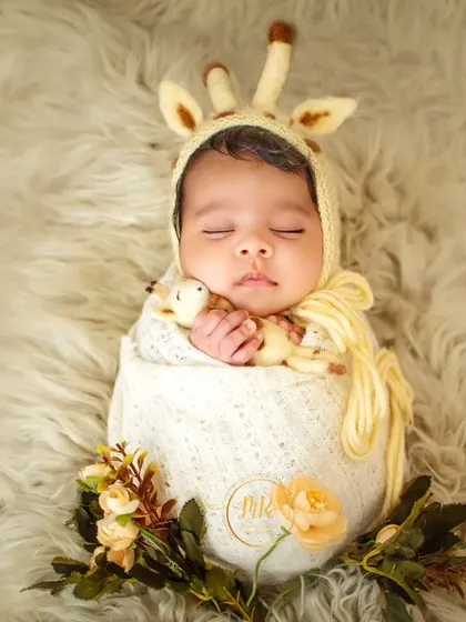 A sleeping newborn wearing an adorable giraffe-themed bonnet, swaddled in a cream-colored wrap on a soft, furry surface.