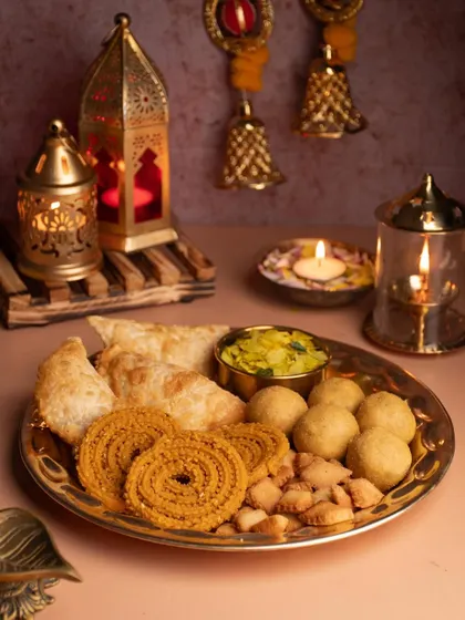A traditional Diwali platter filled with homemade 'Faral'. The warm glow of diyas and lanterns in the background creates a nostalgic and authentic festive atmosphere.