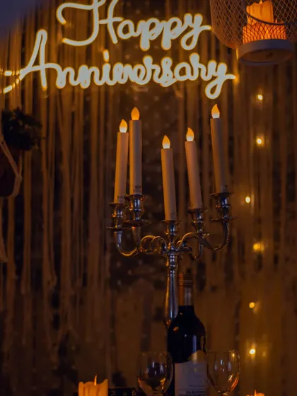 The focus of this anniversary setup is the warm, ambient lighting. A custom "Happy Anniversary" neon sign glows against a macrame backdrop, complemented by a classic candelabra for a timelessly romantic feel.