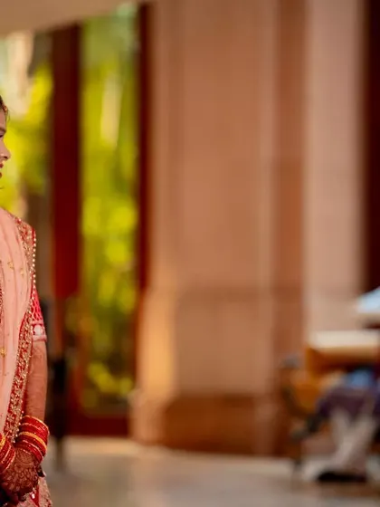 A candid shot of a bride in her red wedding lehenga, looking towards the groom in the background. The composition tells a story of anticipation and connection.