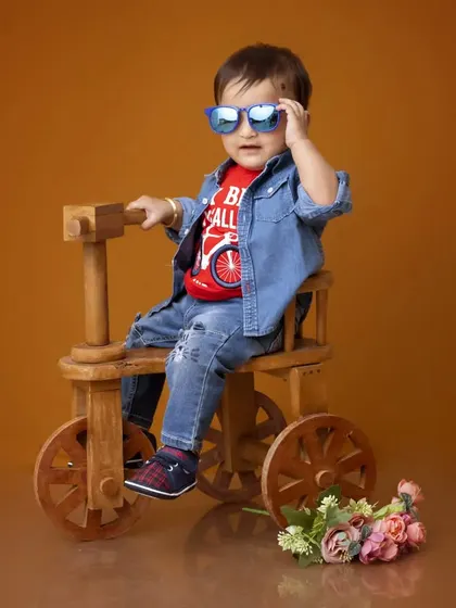 A fun and stylish studio portrait of a young boy for his pre-birthday photoshoot. He is dressed in a denim jacket and sunglasses, posing confidently on a wooden tricycle prop.