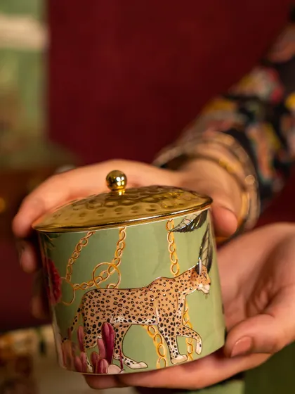 A detail of a jar from the Tales of Tropic collection, held to showcase its fine craftsmanship. The leopard motif and gold lid add a touch of wild sophistication to the vibrant botanical design.