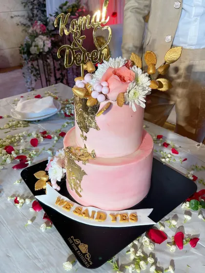 A two-tiered pink engagement cake decorated with gold leaf, white and pink flowers, and a "We Said Yes" banner. This cake was the centerpiece of a beautiful celebration table scattered with flower petals.
