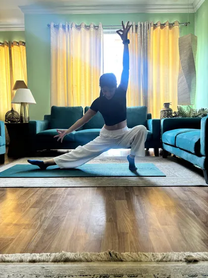 Low lunge with one arm sweeping overhead, sunlight through the curtains behind.