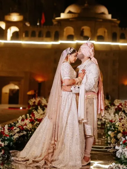 An intimate moment for the couple against the illuminated facade of a Jaipur palace. The wedding aisle is lined with lush red and white florals, creating a romantic pathway for their grand entrance.