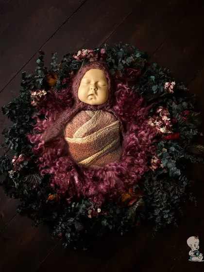 A beautifully composed overhead shot of a newborn wrapped in a textured swaddle, lying in a dark wreath. The contrast between the baby and the dark, moody tones creates a powerful and artistic photograph.