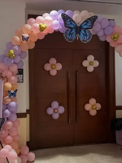 A butterfly-themed balloon arch at an entrance. The arch is made of pastel pink and purple balloons with flower and butterfly accents, welcoming guests to a Mundan and Birthday party.