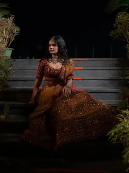 A bride in a stunning rust-colored lehenga, seated on a staircase at night. This portrait is dramatic and elegant, using the ambient light and setting to create a regal mood.