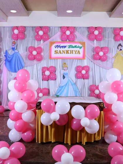 A Cinderella-themed birthday party decoration. The backdrop features Disney characters against a draped background, with pink and white balloon columns and flower-shaped balloon clusters.
