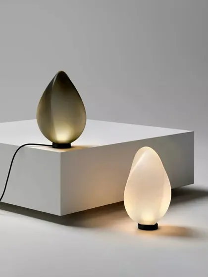 The Solace table lamp by Ross Gardam is presented in two distinct forms, one a luminous white and the other a smoky grey. These sculptural glass pieces, shaped like luminous tears, exemplify my curation of smaller, artistic objects that provide both function and beauty for intimate settings.