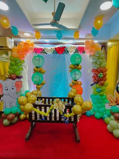A playful jungle themed naming ceremony setup. This decor features a central cradle decorated with garlands, surrounded by a colorful balloon arch and life sized cutouts of cartoon animals like a lion, elephant, and monkey.