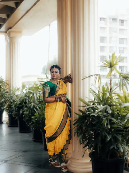 A beautiful portrait of a Maharashtrian bride in her yellow Nauvari saree, standing elegantly amongst pillars and greenery.