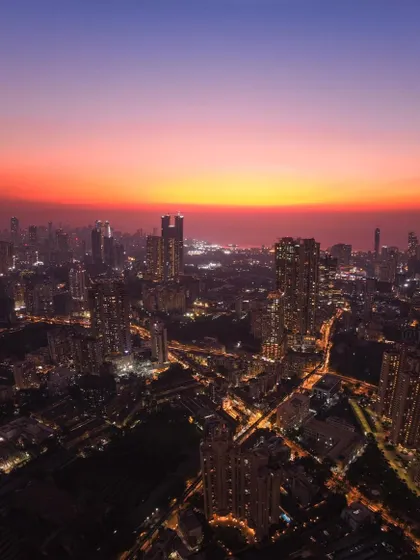 A high-altitude drone shot of Mumbai after sunset, with the city lights beginning to glow under a fiery orange sky.