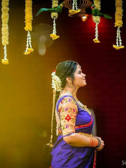 A bride in a beautiful blue saree stands under hanging floral decorations, her profile lit softly.