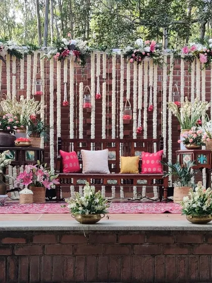 A beautiful rustic Mehendi stage setup against a brick wall. We used hanging strings of tuberose, lush floral arrangements in brass pots, and traditional wooden seating to create a warm and earthy vibe.