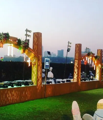 A beautiful buffet setup on a rooftop with a city skyline view. The wooden archways and warm hanging lights create a magical atmosphere for an evening reception or sangeet ceremony.