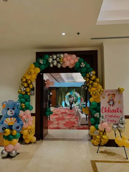 A themed entrance for a child's birthday party. The balloon arch in green, yellow, and pink is complemented by a Care Bear balloon column and a custom welcome sign for 'Chhavi's Birthday Party'.