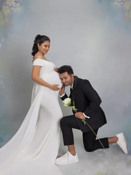 A sweet moment of connection with their unborn twins. The father-to-be kneels to listen to his partner's belly, holding a white rose, set against a dreamy, painted backdrop.