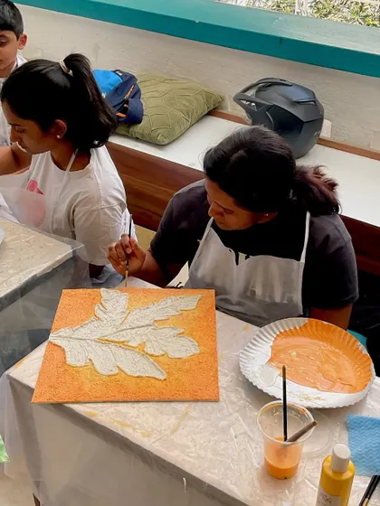 A participant painting her textured leaf canvas. The vibrant orange background makes the green leaves pop.