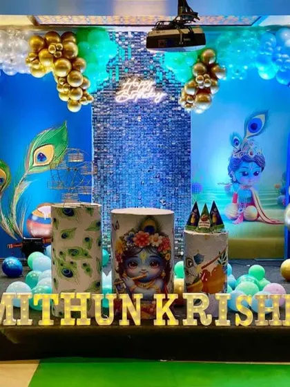 An elaborate Little Krishna themed birthday party setup. This design features custom printed backdrops with Krishna illustrations, peacock feather motifs, themed plinths, and a blue and gold balloon garland, creating a divine atmosphere for the celebration.