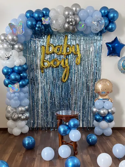 A classic "Baby Boy" welcome home decoration. The setup includes a blue and silver balloon arch over a silver fringe curtain, with a baby-shaped foil balloon accent.