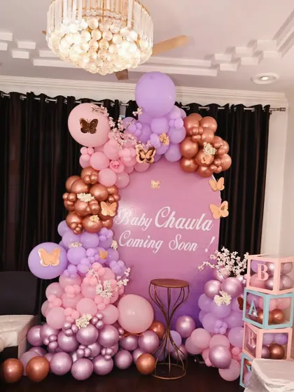 A beautiful butterfly-themed baby shower decoration. This setup uses a soft palette of pink, purple, and rose gold balloons, a personalized "Baby Chawla Coming Soon" backdrop, and charming details like butterfly accents and 'BABY' blocks.