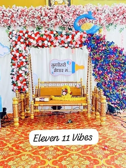 A traditional baby shower setup with a decorated swing as the centerpiece. The backdrop is adorned with a cascade of flowers and a peacock design, complemented by 'BABY' blocks on the side.