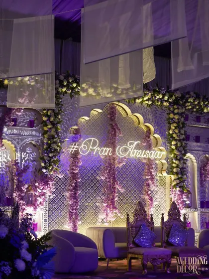 A close-up of the stage, showing the neon hashtag and the beautiful floral garlands hanging against the illuminated jali backdrop. The purple velvet seating adds a touch of royalty.