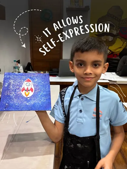 A young boy proudly holds his painting of a rocket ship in a starry galaxy, a perfect example of how art allows for self-expression.