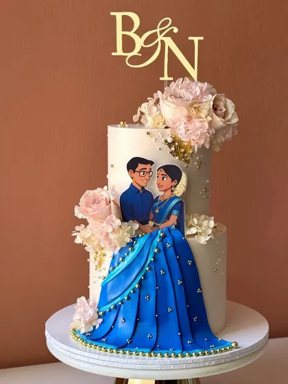 An elegant two-tiered engagement cake featuring a custom illustrated topper of the couple in traditional Indian attire, surrounded by soft pink roses.