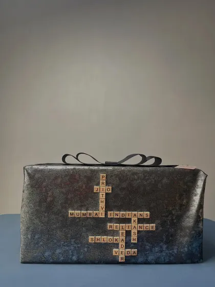A truly personal touch, this gift is wrapped in dark, textured paper and features a custom Scrabble design with the names of family members and important connections, like 'Prithvi', 'Shloka', and 'Mumbai Indians'.