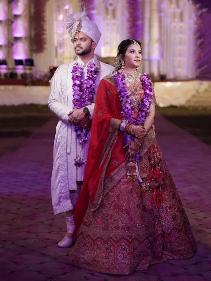 A formal portrait of the bride and groom on their wedding day, adorned with beautiful purple garlands.