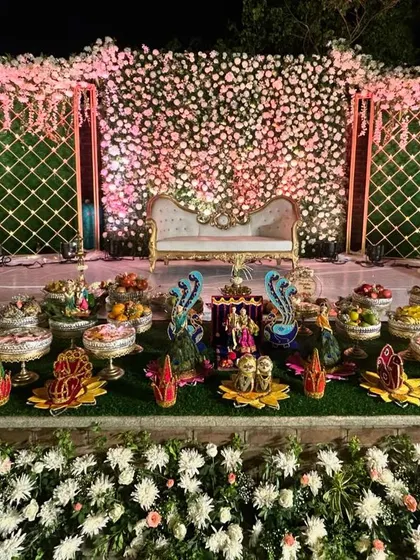 An engagement stage decorated with a wall of flowers and traditional offerings, beautifully lit for an evening ceremony.