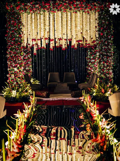A dramatic wedding stage setup with a mirrored aisle reflecting the floral mandap. The combination of traditional flowers like jasmine and roses with modern reflective surfaces creates a visually stunning effect, perfect for a memorable evening reception.
