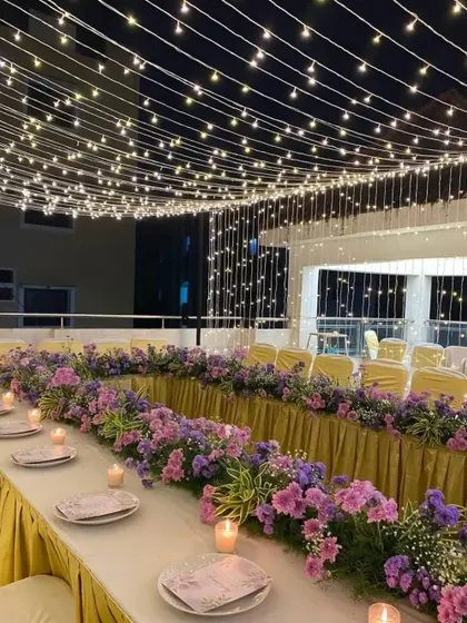 An elegant dinner setup under a canopy of fairy lights. The long table is decorated with a beautiful floral runner and candles, creating a magical and intimate dining experience.