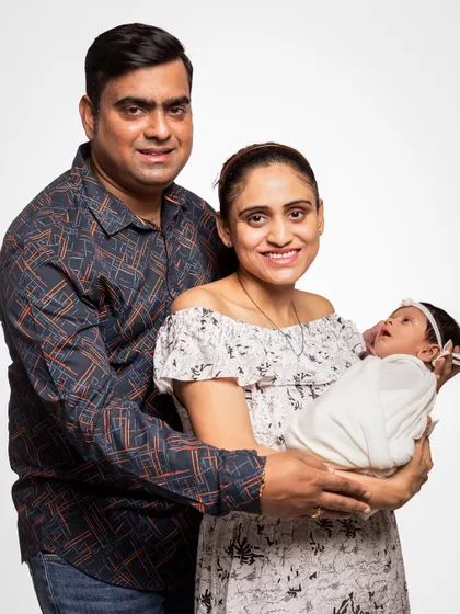 A classic family pose celebrating your new arrival. This portrait features both parents smiling for the camera while holding their swaddled newborn, a perfect memory of when three became one.