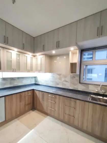 This simple and functional L-shaped kitchen is designed for modest living. The dual-tone laminate cabinets in light wood and grey offer ample storage, while under-cabinet lighting ensures a well-lit workspace.
