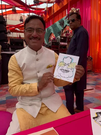 A smiling guest with his caricature at a wedding event. The colorful pandal in the background adds to the festive atmosphere.