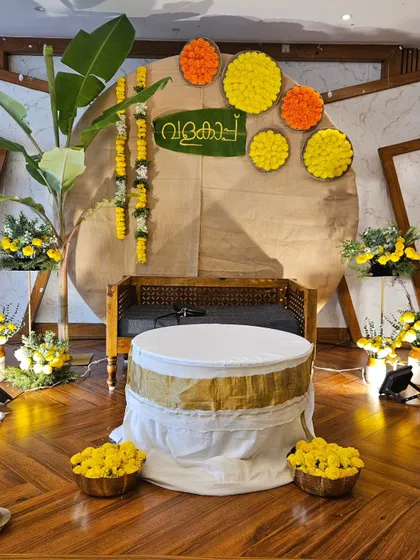 A complete view of the traditional Valaikappu stage. The setup includes a comfortable wooden bench for the mom-to-be, lit by traditional brass Nilavilakku lamps, and surrounded by bowls of fresh marigold flowers to complete the auspicious setting.