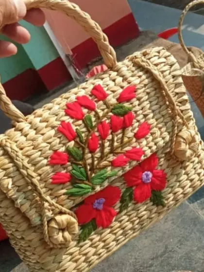 This rectangular handbag is woven from thick natural fibers and features vibrant red floral embroidery. Its structured shape and braided handles make it a sturdy and stylish choice for everyday use.