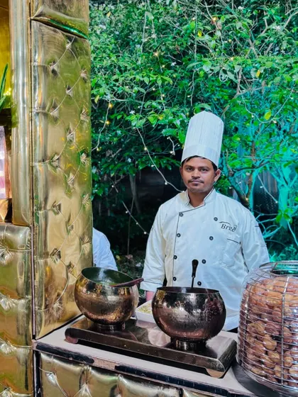 A chef at the pani puri section of the chaat counter, set against a lush green backdrop.