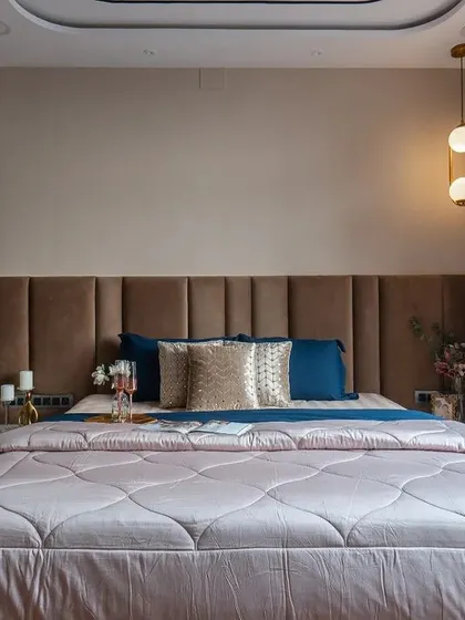 A classic bedroom interior with a plush brown headboard, elegant patterned curtains, and a beautifully designed false ceiling. The warm lighting creates a cozy and inviting atmosphere.
