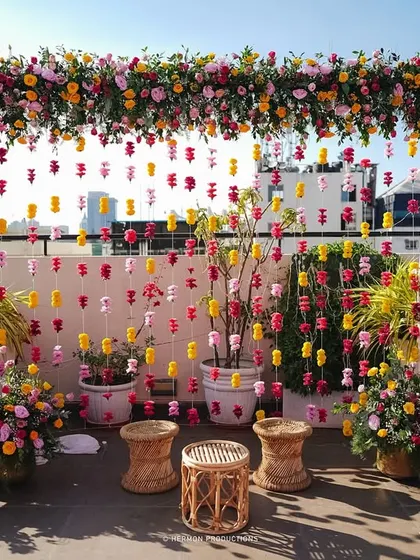 A vibrant rooftop Mehendi setup with a backdrop of hanging flowers in shades of pink and yellow, perfect for an urban celebration.