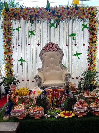 A unique baby shower decoration featuring a floral arch with hanging parrot motifs for a touch of nature and tradition. The seer plates are filled with fruits, sweets, and flanked by traditionally dressed dolls.
