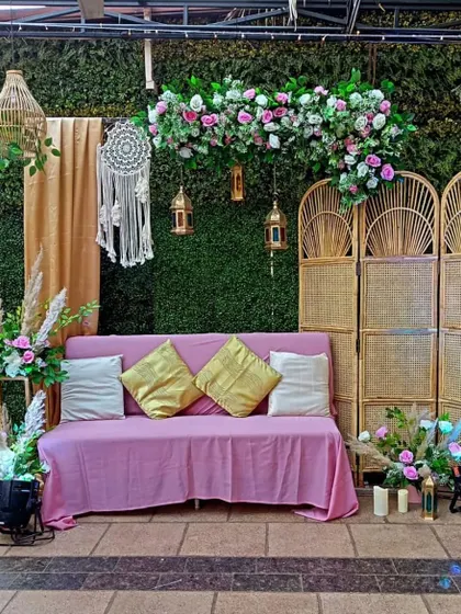 A cozy and chic boho-themed seating area for a relaxed party. The setup includes a green hedge wall, a dreamcatcher, and comfortable seating with plush cushions.