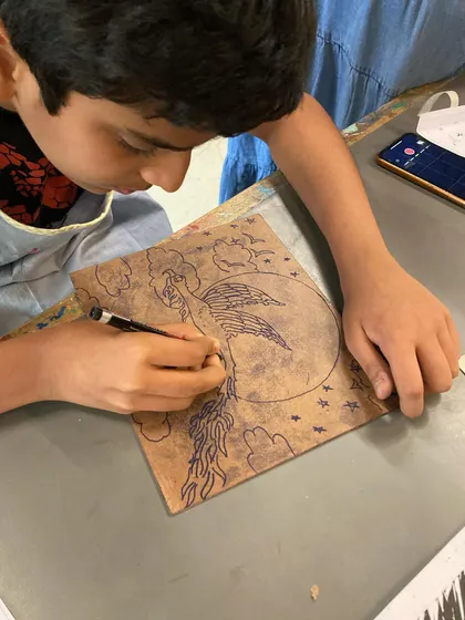 A young artist concentrates on drawing his phoenix design directly onto a block. This is the first step in creating a relief print, where every line he draws will soon be carved to create a stunning image.
