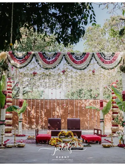 A beautiful mandap setup where tradition is woven into every element. The stage is framed by pillars wrapped in banana leaves and floral garlands, with a backdrop of white chrysanthemums, nerium, and hanging temple bells.