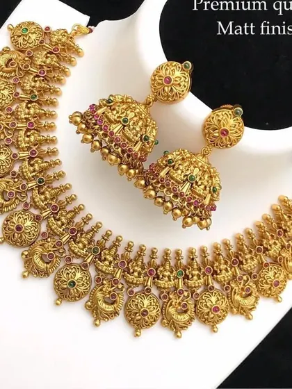 A beautiful temple necklace set with matching jhumkas. The design features tiny goddess figures and peacock motifs with red and green stones, all in a premium matte finish.