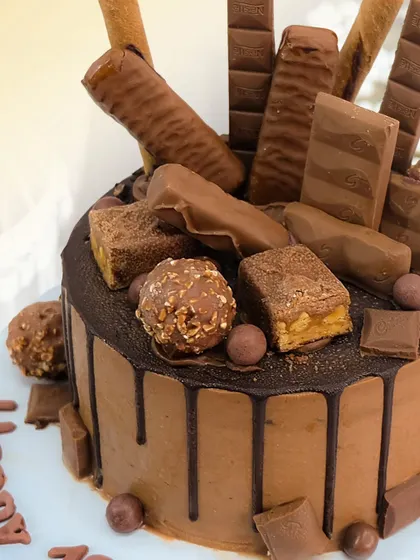 A close-up of the Chocolate Overload cake, showing the rich drip and the generous pile of assorted chocolate bars and treats.